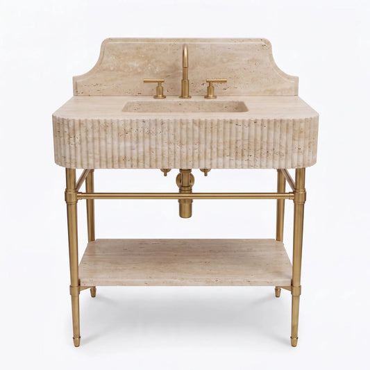 A freestanding bathroom vanity with a beige stone basin and backplate, fluted front, brass faucet and handles, and a lower shelf on a light gray background.