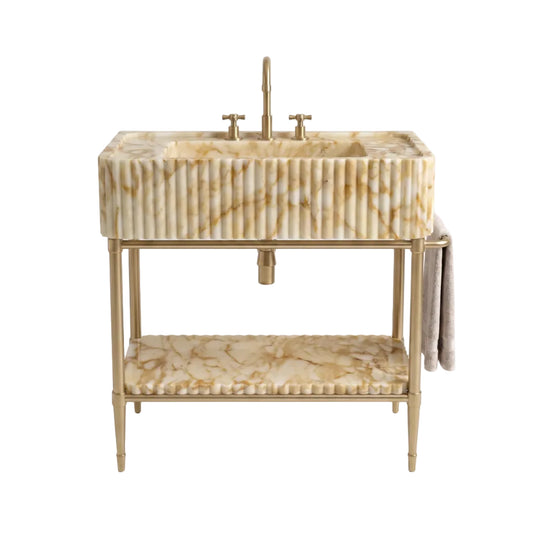 A freestanding vanity sink with a rectangular fluted marble basin in warm cream and tan tones, a matching lower shelf, and a gold-tone metal frame and faucet, shown on a clean white background.