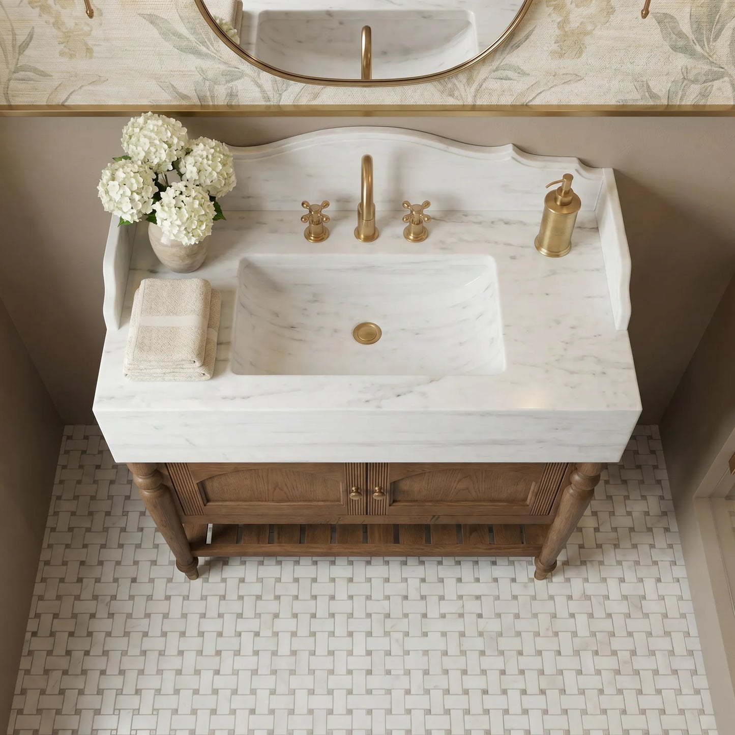 A bathroom vanity featuring a white marble sink countertop with a warm brass faucet, handles, and soap dispenser, mounted on a rustic brown wooden cabinet. The scene is styled with white flowers and a folded towel, set against a beige wall and a woven tile floor.