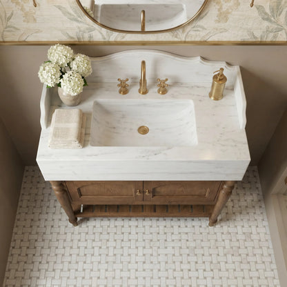 A bathroom vanity featuring a white marble sink countertop with a warm brass faucet, handles, and soap dispenser, mounted on a rustic brown wooden cabinet. The scene is styled with white flowers and a folded towel, set against a beige wall and a woven tile floor.